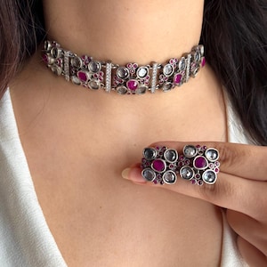 May include: A silver-toned choker necklace and matching earrings set. The jewelry features alternating clear and ruby-colored stones, with small clear stones between the larger stones. The necklace is around the neck.