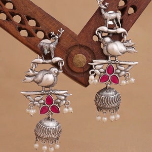 May include: Silver tone earrings with a bird, deer, and red stone accents. The earrings have a dangling bell charm with white pearl accents.