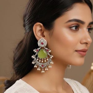 May include: Silver-toned statement earrings with a pear-shaped green stone, red accents, and small white pearls. The earrings have an ornate design with a floral motif and are worn by a person with dark hair.