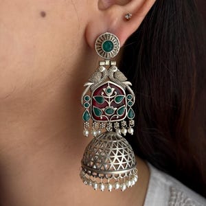 May include: Silver-toned dangle earrings with a detailed design. The earrings feature a circular top with a green stone, two bird figures, and a red and green floral design. The bottom has a bell shape with a geometric pattern and small white beads.