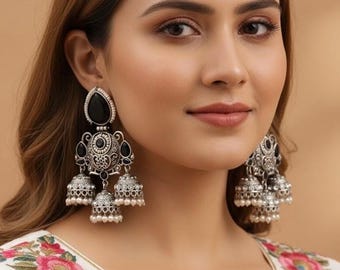 May include: Silver-toned chandelier earrings with black teardrop-shaped stones and small white pearl accents. The earrings feature intricate filigree designs and three dangling bell-shaped elements. The earrings are worn by a person wearing a white top with floral embroidery.