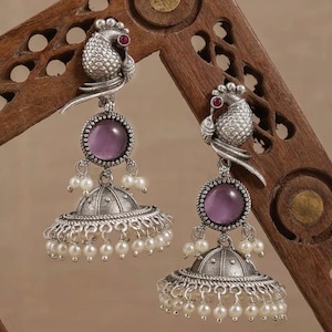 May include: Silver tone earrings with a purple stone and a peacock design. The earrings have a dangling bell shape with pearl accents.