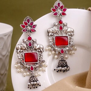 Può includere: Orecchini color argento con un centro di pietre preziose rosse e un design floreale nella parte superiore. Gli orecchini presentano due figure di elefanti e piccole perle bianche. La parte inferiore degli orecchini ha un design a forma di campana con più perle.
