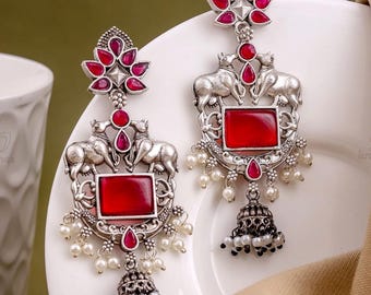 May include: Silver-toned earrings with a red gemstone center and a floral design at the top. The earrings feature two elephant figures and small white pearls. The bottom of the earrings have a bell-shaped design with more pearls.