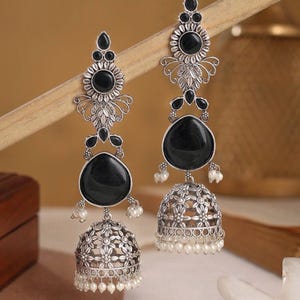 May include: A pair of silver-toned dangle earrings with black gemstone accents and small white pearls. The earrings feature a floral design and a bell-shaped bottom. The earrings are displayed against a neutral background.