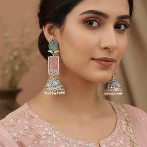 May include: Silver-toned earrings with a teardrop-shaped green stone, a pink rectangular stone, and a bell-shaped bottom. The earrings have intricate detailing and small white beads. The woman in the image is wearing a pink embroidered top.