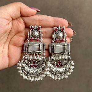 May include: A pair of silver earrings with intricate designs, featuring a crescent moon shape, a rectangular mirror, and red gemstones. The earrings are adorned with white pearls.