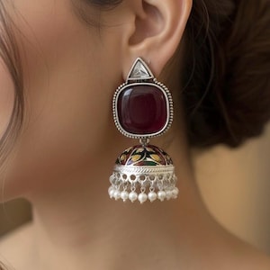 May include: Silver earrings featuring a large, square, burgundy stone and a small, triangular, clear stone. The lower section is bell-shaped, adorned with colourful floral patterns and small white pearl dangles. The earrings are worn on an ear.