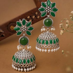 May include: A pair of silver-toned earrings with green gemstone accents. Each earring features a flower-shaped top, a round green stone, and a bell-shaped bottom adorned with green stones and small white pearls. The earrings have a vintage aesthetic.