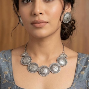 May include: A silver-coloured jewellery set comprising a necklace and matching earrings. The necklace features circular pendants with a floral design. The earrings are round and have a similar floral pattern. The jewellery is worn against a grey top.