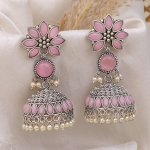 May include: A pair of pink and silver-toned jhumka earrings. The earrings feature a floral design at the top, a round pink stone, and a bell-shaped bottom with pink accents and dangling pearls. The earrings are set against a white background.