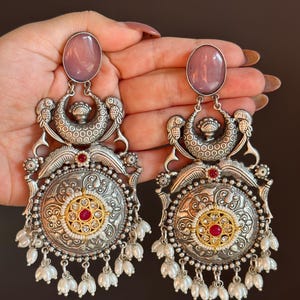 Puede incluir: Un par de pendientes de plata con una piedra rosa, una gota de perla y diseños florales intrincados.