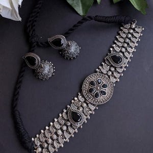 May include: A silver-toned choker necklace and matching earrings set with black gemstone accents. The necklace features a central circular pendant and teardrop-shaped stones. The earrings have a teardrop-shaped black stone and a round, textured base.