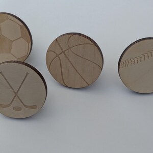 Op de afbeelding: Vier houten decoratieve knopen met sportthema's. Ontwerpen omvatten een voetbal, basketbal, honkbal en gekruiste hockeysticks. De knopen zijn lichtbruin met geëtste details, perfect voor knutselen of een sportieve touch.