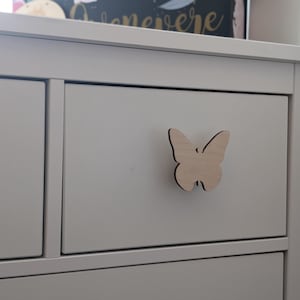 May include: White chest of drawers with three drawers. The top drawer has a wooden butterfly-shaped knob.