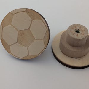 Op de afbeelding: Twee houten kastknoppen. Eén is rond met een voetbalontwerp, met lichtgekleurde vijfhoeken op een donkerdere houten achtergrond. De andere knop heeft een gelaagde, cilindrische vorm met een schroefinzet.