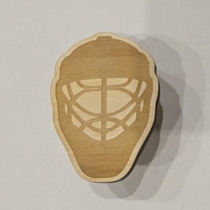 May include: A wooden hockey goalie mask cutout. The mask is light brown with a darker brown outline. The mask has a traditional cage design.
