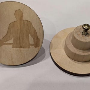 Könnte beinhalten: Flaschenverschluss aus Holz in Form eines Hockeyscheibens mit einer Silhouette eines Hockeyspielers. Der Verschluss hat eine runde Basis und eine mehrstufige Holzoberseite mit einer Schraube. Der Puck hat einen dunklen Rand.