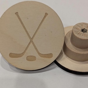 Könnte beinhalten: Ein runder Holzscheibe mit einem geschnitzten Design aus Hockeyschläger und Puck. Die Scheibe ist mit einer Schraube an einer Holzbasis befestigt.
