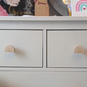 May include: White chest of drawers with two drawers featuring sun-shaped wooden knobs. The drawers are closed.