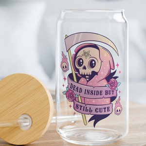 A glass can with a pink and purple graphic of a cartoon skeleton wearing a hooded cloak. The text on the graphic reads 'Dead inside but still cute.' The can has a bamboo lid and a straw.