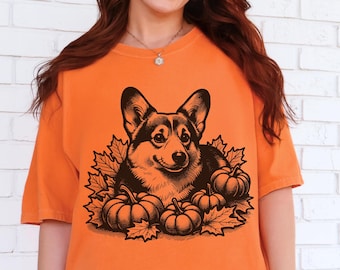 May include: A burnt orange short-sleeve t-shirt featuring a black graphic print of a Corgi dog surrounded by autumn maple leaves and pumpkins. The design is centered on the chest of the garment, which has a crew neckline and a relaxed fit.