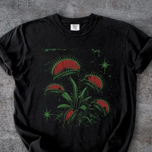 May include: A black short-sleeve t-shirt featuring a graphic print of a Venus flytrap plant with red interior lobes and green stems. The design includes small green star-like accents and a distressed texture. The shirt has a crew neckline.