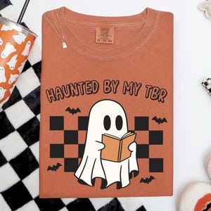 An orange shirt featuring a ghost reading a book, bats and a retro checkered pattern in the background. Text reads haunted by my tbr. Tbr means to be read.