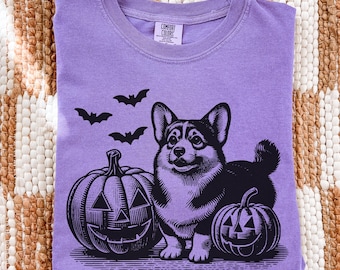 May include: A purple Comfort Colors t-shirt featuring a Halloween design. The graphic includes a black and white corgi dog, two jack-o'-lanterns, and three bats. The shirt is folded neatly on a textured, neutral-colored surface.