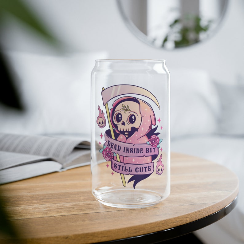 A clear glass can-shaped drinking glass with a cartoon grim reaper design. The reaper is wearing a pink hooded robe and holding a scythe. The text "DEAD INSIDE BUT STILL CUTE" is on a banner below the reaper.