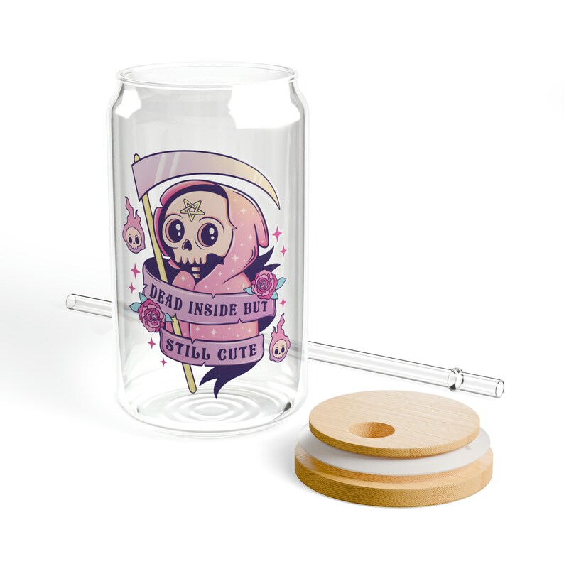 A clear glass can-shaped tumbler with a bamboo lid and a glass straw. The tumbler features a cartoon grim reaper design with the text "DEAD INSIDE BUT STILL CUTE" on a banner. The design includes pink, purple, and white accents.