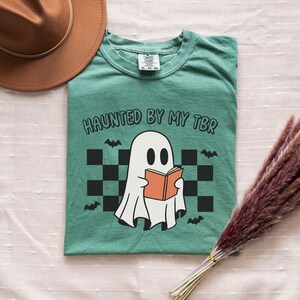 A ghost reading a book, bats and a retro pattern in the background. Text reads haunted by my tbr.