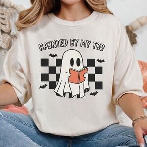 Halloween t-shirt for book readers, featuring a ghost reading a book, bats and a retro pattern in the background. Text reads haunted by my tbr.