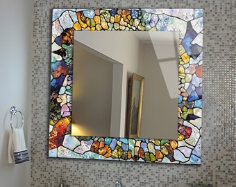 May include: Square bathroom mirror with a colorful, stained-glass style border. The border features a mosaic of blues, greens, yellows, and reds. The mirror is mounted on a tiled wall above a sink.