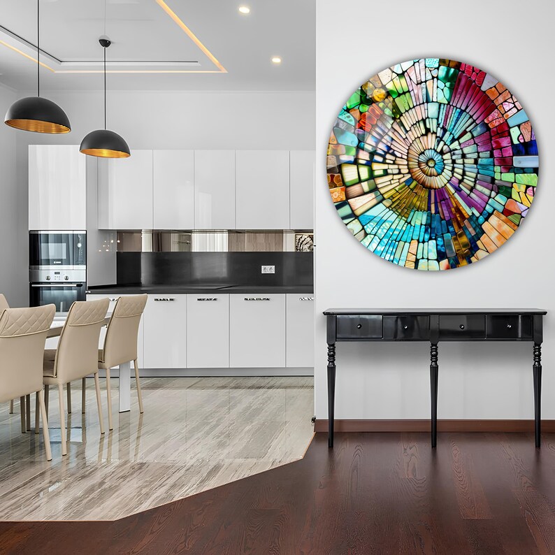 Op de afbeelding: Een kleurrijk rond moza&iuml;ek wandkunstwerk met een spiraalvormig ontwerp. De kunst hangt aan een witte muur in een moderne keuken met een zwart aanrechtblad en witte kasten.