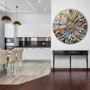 May include: A colorful abstract circular wall art piece with a geometric design. The art is hanging on a white wall above a black console table with three drawers. The table has four legs and is in a modern dining room with a white kitchen in the background.