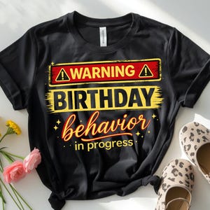 May include: Black t-shirt featuring a distressed yellow and red graphic that reads "WARNING BIRTHDAY behaviour in progress." The shirt has a crew neck and short sleeves. Leopard print shoes are visible.
