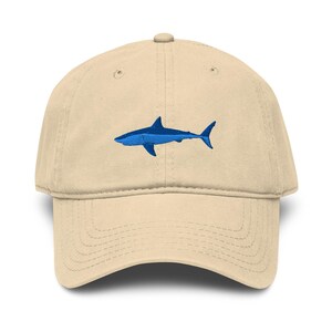 Puede incluir: Gorra de béisbol beige con un diseño de tiburón azul bordado. La gorra tiene una visera curva y una correa ajustable en la parte trasera, ofreciendo un look casual y deportivo.