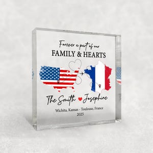 May include: A clear acrylic block featuring a design with the text "Forever a part of our FAMILY & HEARTS." The design includes the USA and France flags, connected by a dotted line with a heart and airplane. The names "The Smith Josephine" and locations are also included.