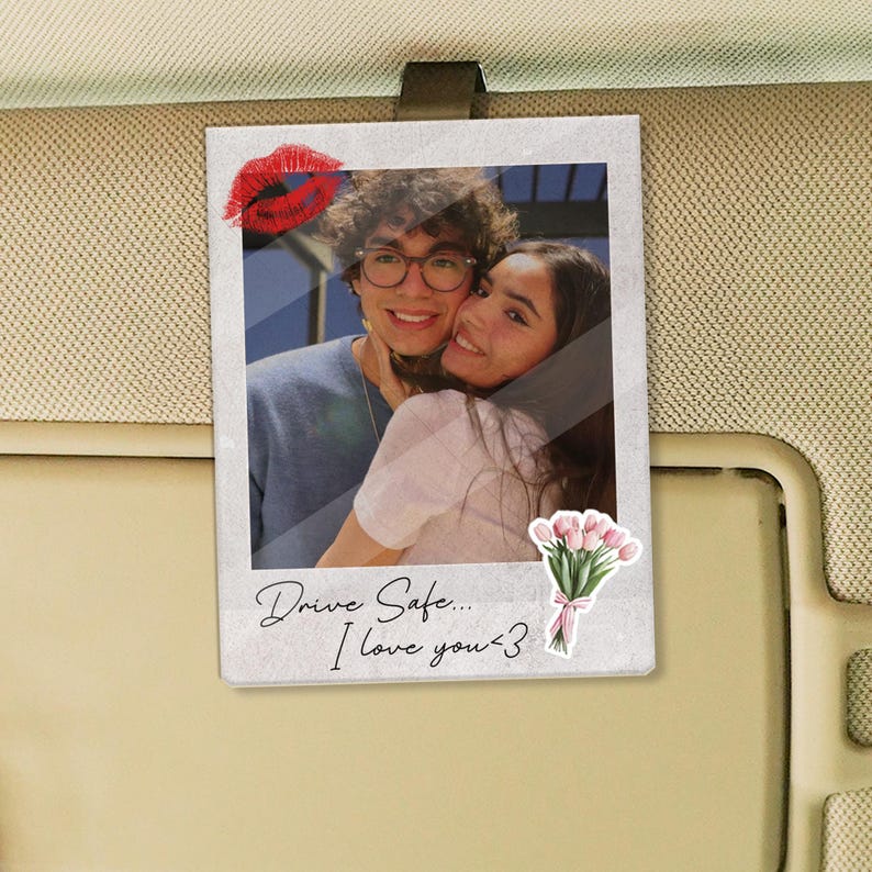 May include: A photo print featuring a couple embracing, with a red lip sticker and a bouquet of pink tulips. The print has the text "Drive Safe... I love you <3". The photo is clipped to a car visor.