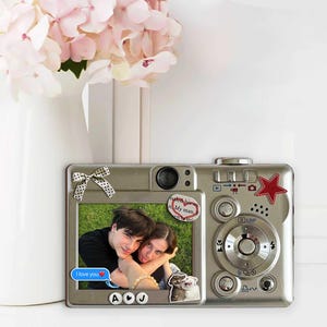 May include: Silver-colored picture frame in the shape of a camera, with a photo of a couple. The frame has decorative stickers, including a red star and a bow. The photo frame is next to a white vase with pink flowers.