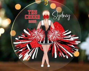 May include: A round clear acrylic ornament featuring a blonde cheerleader in a black and white uniform with a red bow, holding red and white pom-poms. The text reads THS CHEER 2025 and Sydney. The design includes a gold hanging cord at the top.