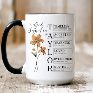 May include: White ceramic mug with a black handle and rim. The mug features the name "Taylor" in large letters, with floral accents and inspirational text including "God Says I Am" and Bible verses. The mug is approximately 4 inches tall.