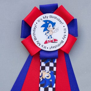K&ouml;nnte beinhalten: Eine festliche Geburtstagsrosette mit dem Charakter Sonic the Hedgehog. Die Rosette hat rote und blaue B&auml;nder, mit einem zentralen runden Emblem mit der Aufschrift "It's My Birthday!". Ein kariertes Band mit Sonics Bild ist ebenfalls vorhanden.