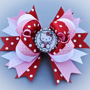 May include: A large, decorative hair bow featuring red and pink ribbons with white polka dots. The bow has a central embellishment with a Hello Kitty image. Accents include white glitter and a pink and white polka dot ribbon.
