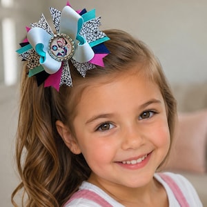 Puede incluir: Un lazo para el cabello colorido con un dise&ntilde;o central con una imagen de personaje, rodeado de capas de cintas blancas, turquesas, azul marino y rosas. El lazo est&aacute; sujeto al cabello de una ni&ntilde;a.