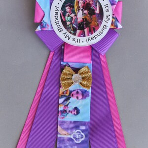 May include: A birthday ribbon in shades of purple, pink, and blue, featuring a circular image of three people. The text "It's My Birthday!" is printed around the image. A gold glitter bow is attached to the ribbon, adding a touch of sparkle.