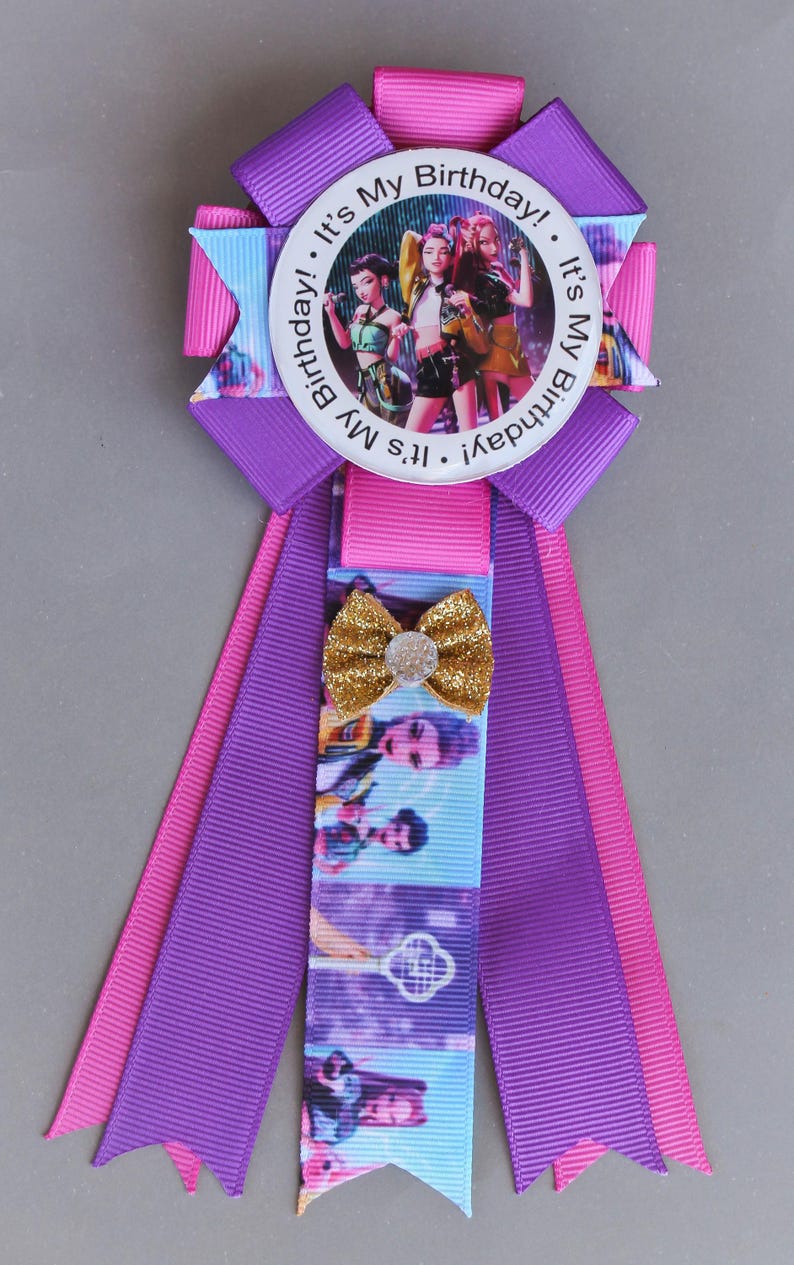 May include: A birthday ribbon in shades of purple, pink, and blue, featuring a circular image of three people. The text "It's My Birthday!" is printed around the image. A gold glitter bow is attached to the ribbon, adding a touch of sparkle.