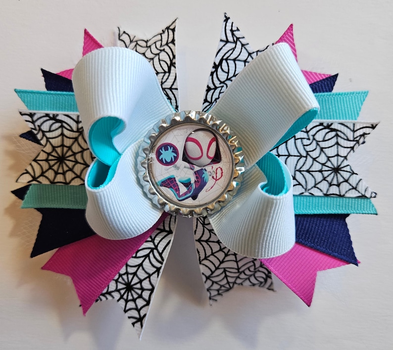 Puede incluir: Un lazo para el cabello hecho a mano con una cinta azul claro con bordes turquesa, superpuesto con cintas rosas, azul marino y turquesa. El lazo tiene un adorno central con un dise&ntilde;o de Spider-Gwen y est&aacute; acentuado con cinta de telara&ntilde;a en blanco y negro.