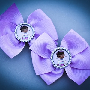 May include: Two lavender hair bows. Each bow features a circular embellishment with a cartoon image of a girl and the text "Doc". The bows are made of a ribbed fabric and are arranged on a blue surface.